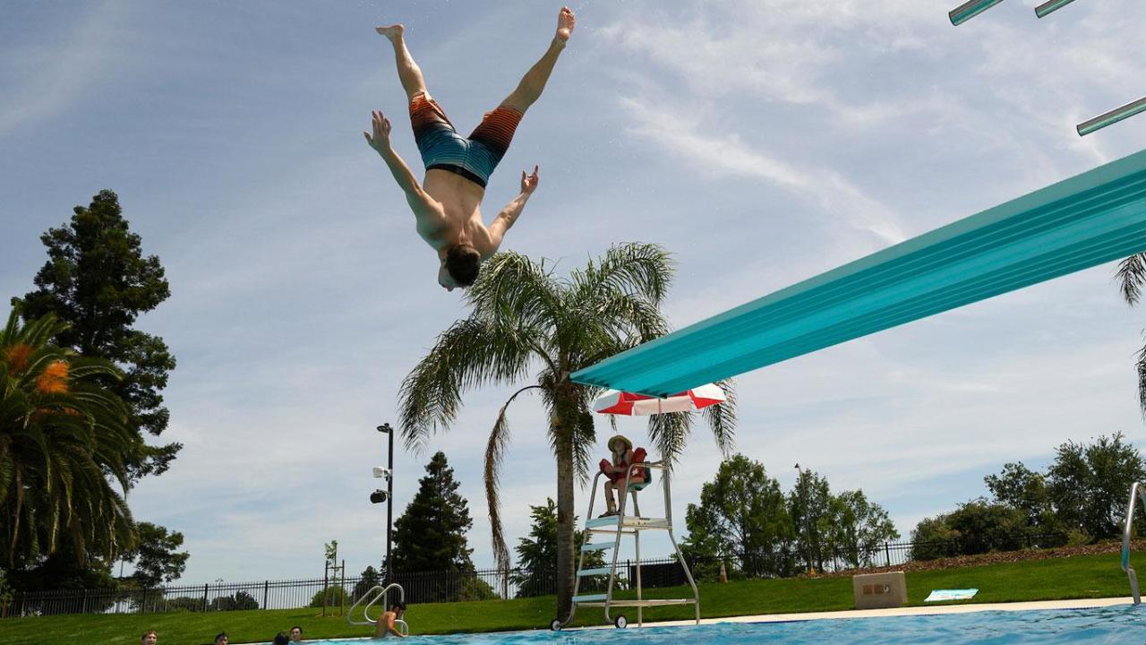 young man doing a flip off a diving board into a pool with lifeguard and palm trees in the background